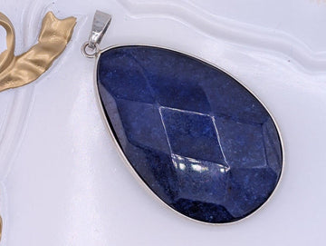 A large, blue gemstone pendant with a silver frame is displayed on a white surface.