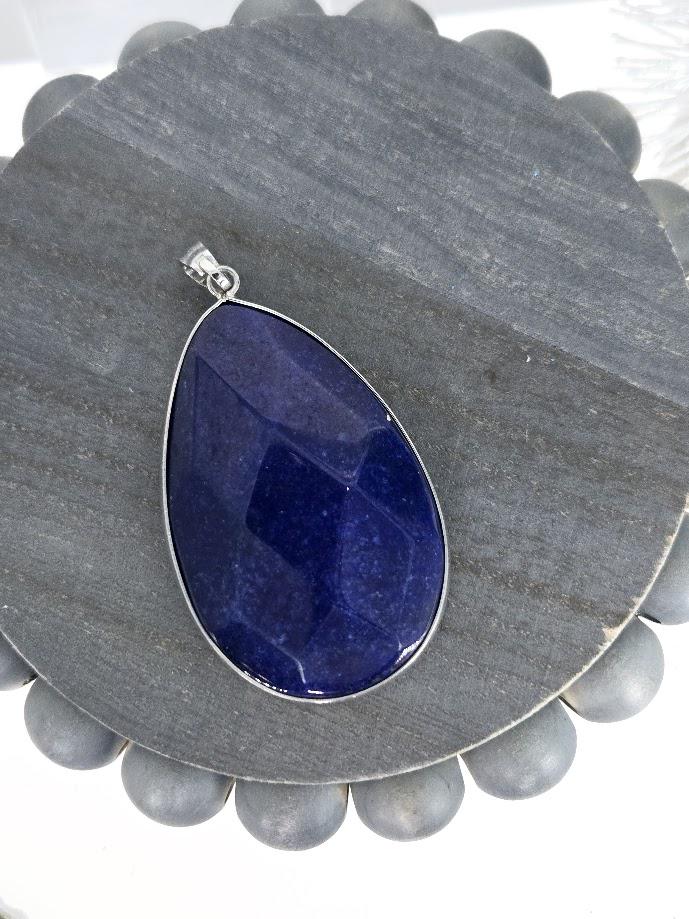 A large, blue gemstone pendant with a silver frame is displayed on a gray wooden surface.