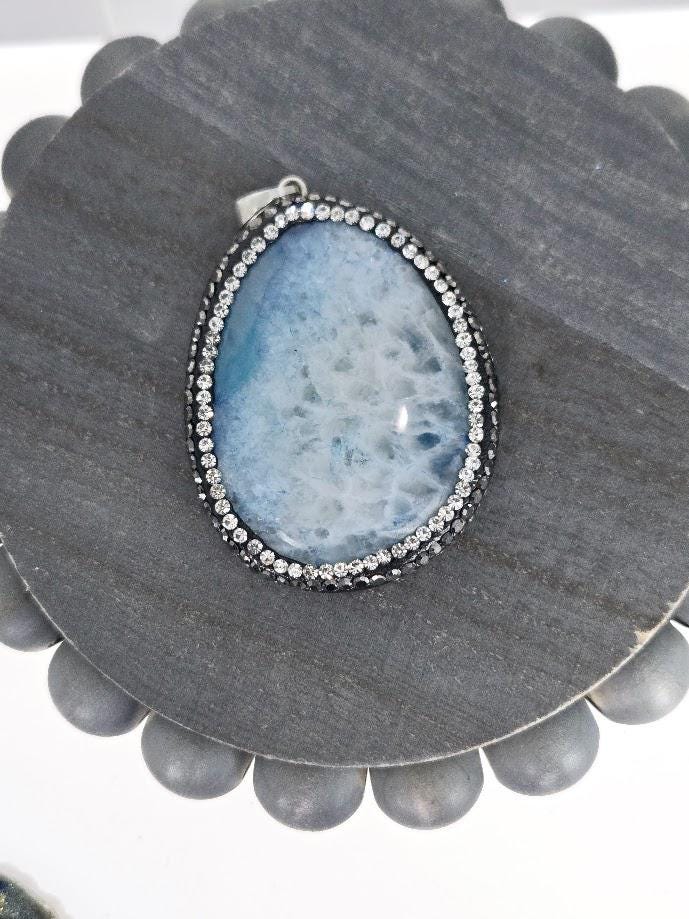 A large, blue-green stone pendant with a silver frame and sparkling rhinestones, resting on a white surface.