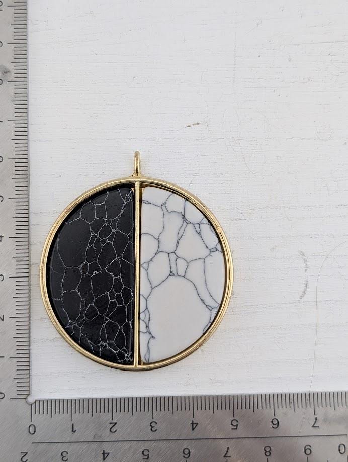 A gold-colored pendant with a black and white marble-like stone in the center, placed on a white surface next to a ruler for size reference.