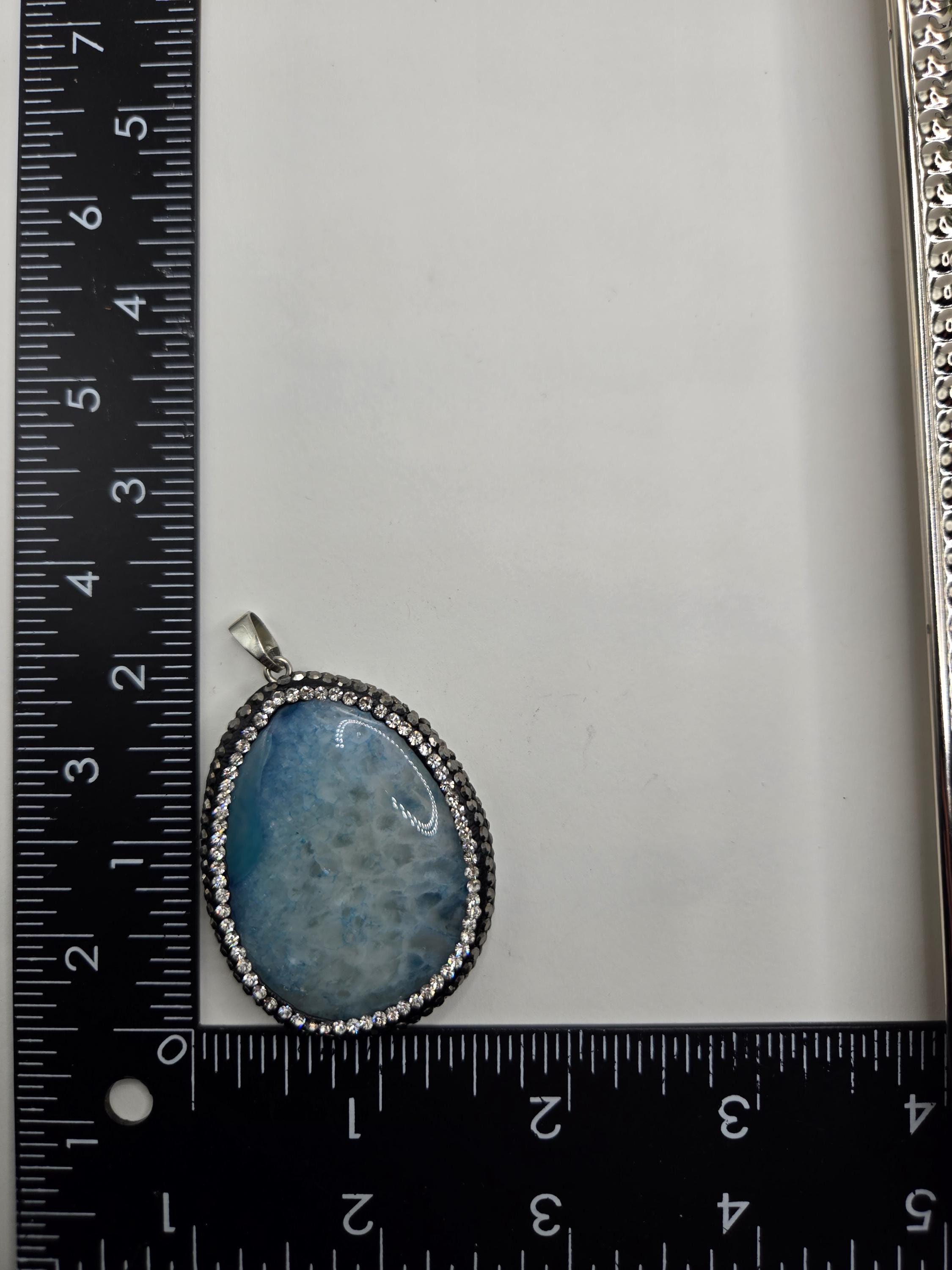 A pendant with a blue stone and a silver chain is placed next to a ruler on a white surface.