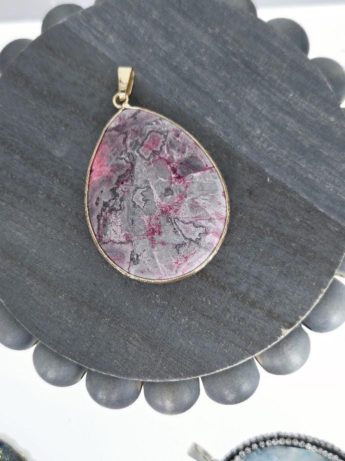 A large, oval-shaped pendant made of a dark-colored stone, possibly jade, with a gold-colored frame. The pendant is hanging from a gold-colored chain.