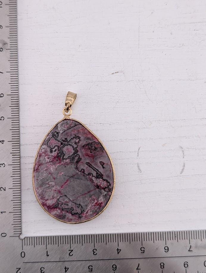 a pendant with a large, oval-shaped stone in a gold frame, placed next to a ruler for size comparison.