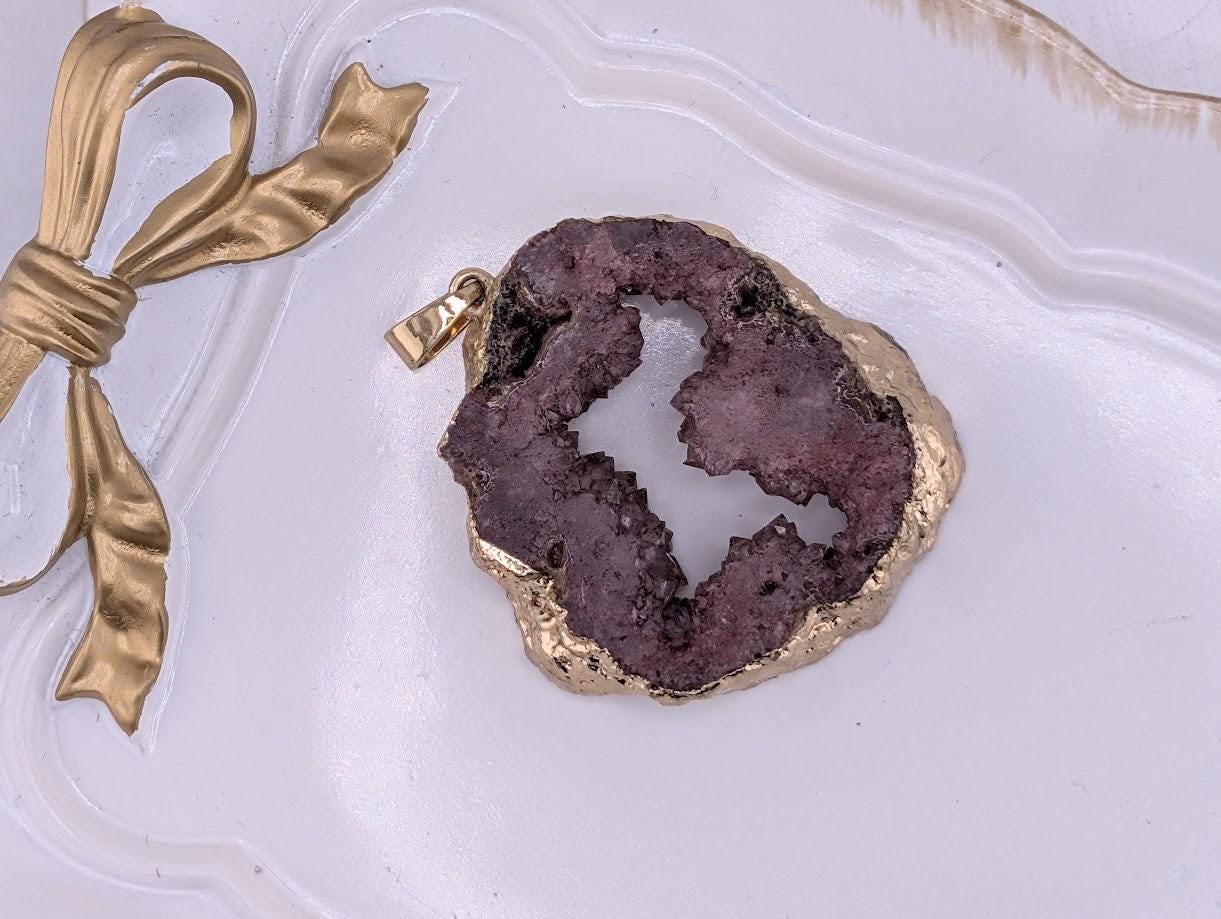 A pendant made of a purple gemstone is displayed on a white surface, accompanied by a gold ribbon.