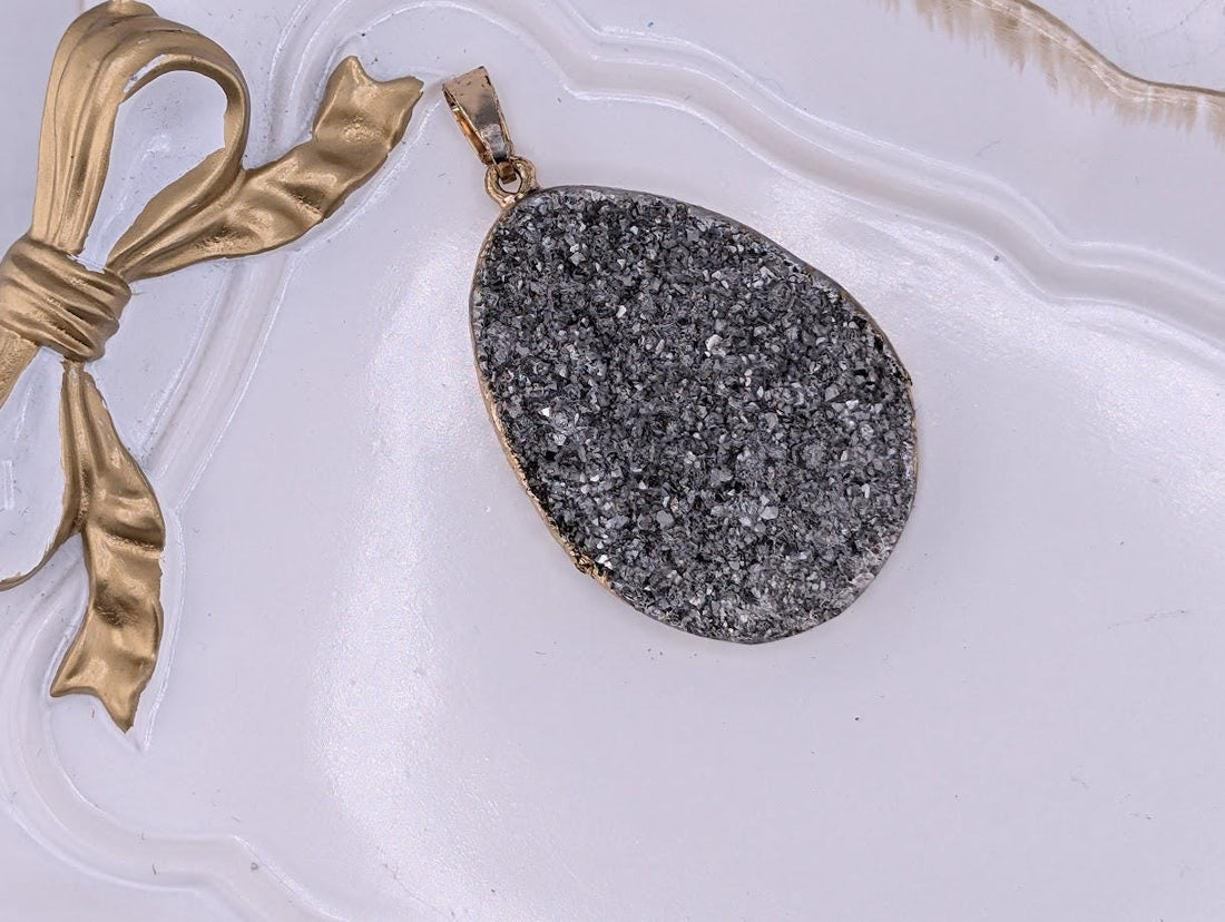 A large, oval-shaped pendant made of gray and black stones is displayed on a white surface, accompanied by a gold ribbon.