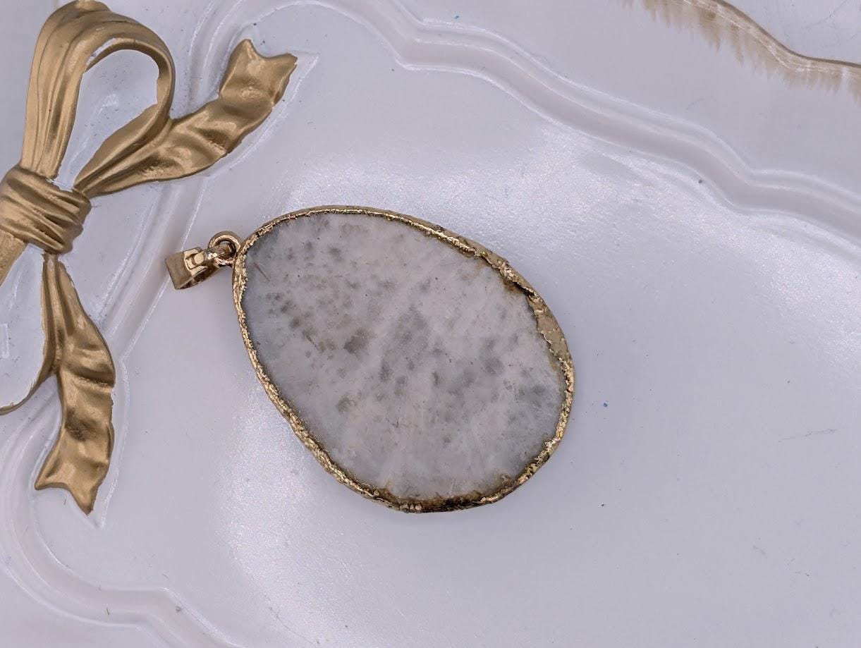 A large, white, and gold pendant with a gold-colored frame is displayed on a white surface, accompanied by a gold ribbon.