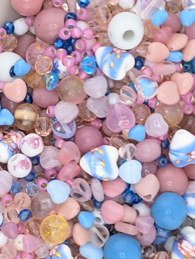 The image is a colorful assortment of various beads, including glass beads in shades of pink, blue, and white, as well as glass beads with intricate designs. The beads are scattered throughout the frame, creating a visually appealing and vibrant display.