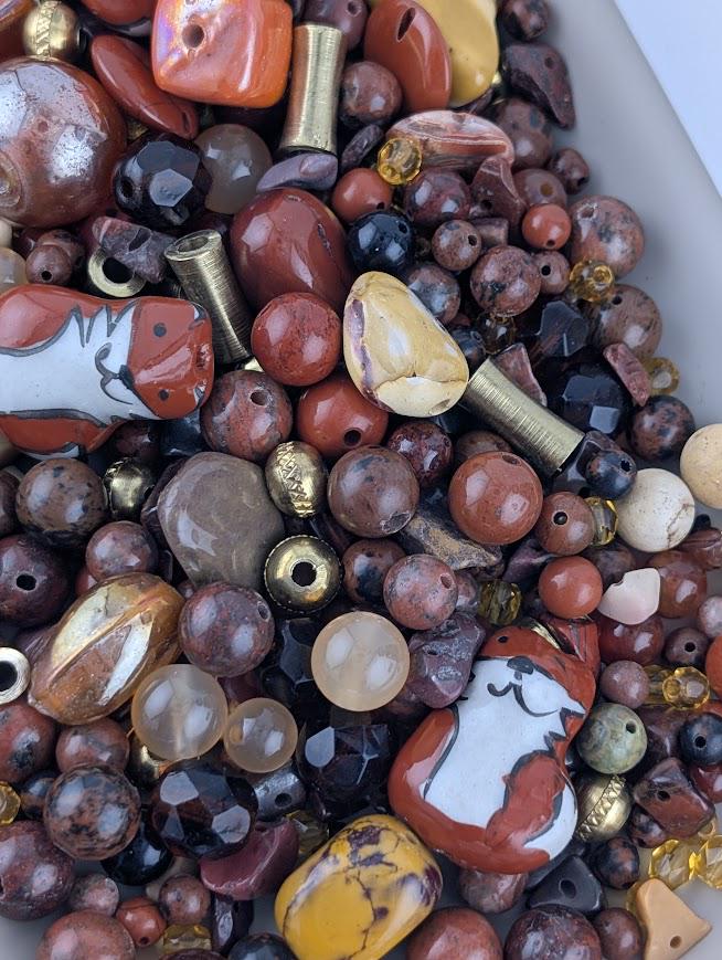The image is a close-up of a pile of various beads and stones, including glass beads, metal beads, and natural stones in different colors and shapes.