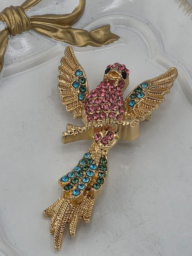 A decorative gold-colored pin or brooch in the shape of a bird, adorned with colorful gemstones.