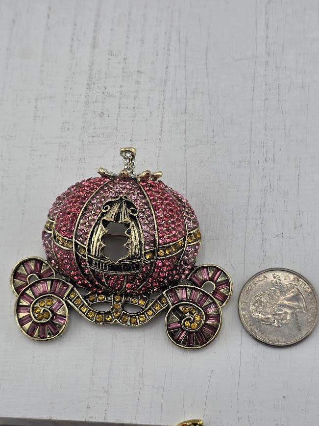 A large, ornate, and intricately designed pendant in the shape of a carriage, adorned with sparkling jewels and gold accents, is placed next to a coin.