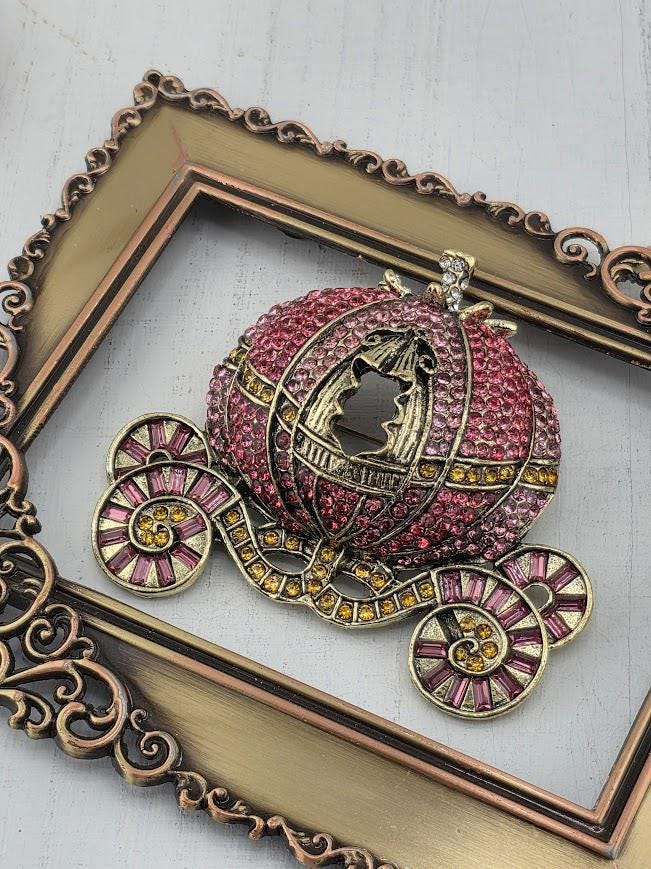 A decorative brooch in the shape of a carriage, adorned with sparkling rhinestones and gold accents, is displayed within an ornate gold frame.