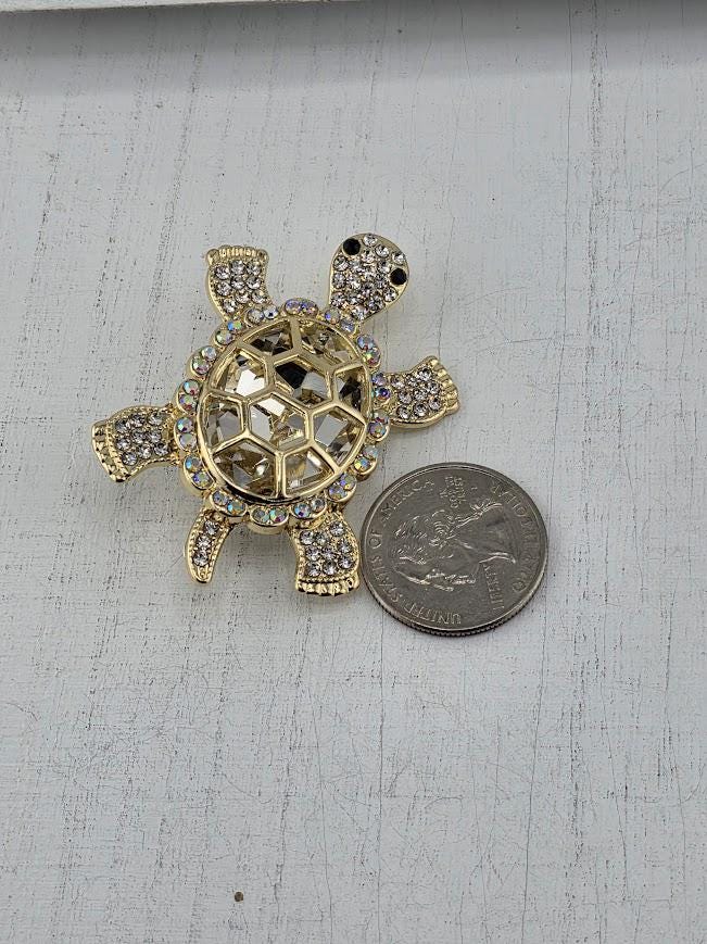 A gold-colored turtle brooch with rhinestones is placed next to a silver coin on a white surface.