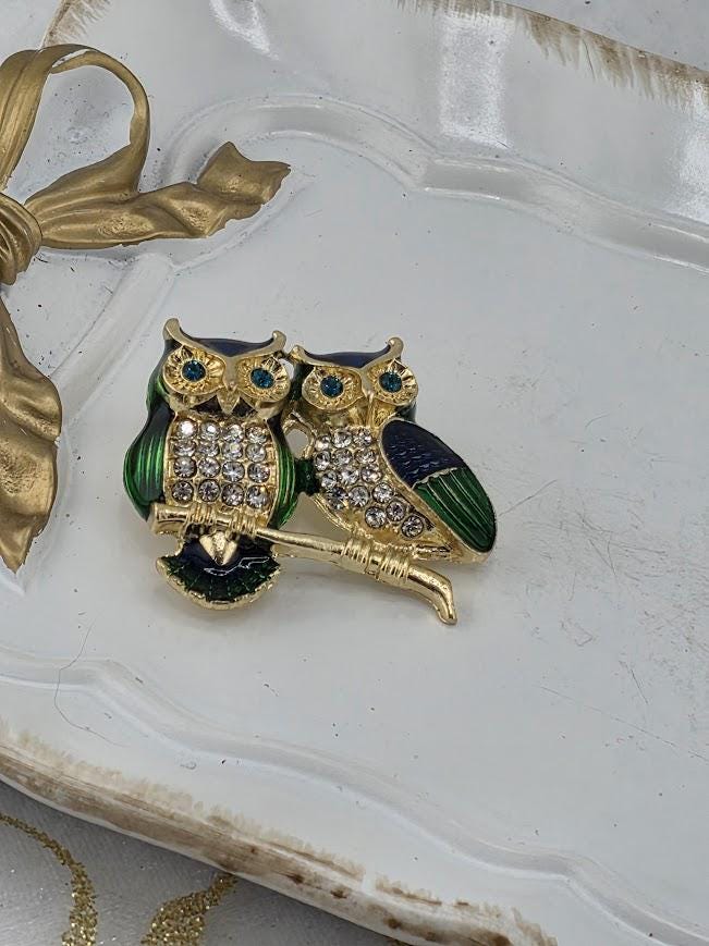 A decorative brooch featuring two ornate owls, one green and one blue, with sparkling gemstones. The owls are perched on a branch-like structure, and the brooch is placed on a white surface.