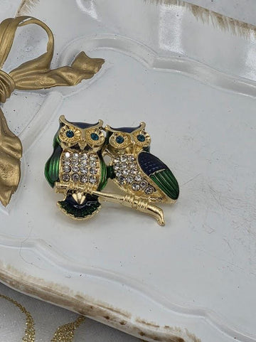 A decorative brooch featuring two ornate owls, one green and one blue, with sparkling gemstones. The owls are perched on a branch-like structure, and the brooch is placed on a white surface.