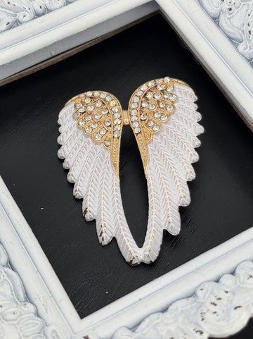 A decorative white and gold wing brooch with rhinestones is displayed against a black background, framed by a white ornate frame.