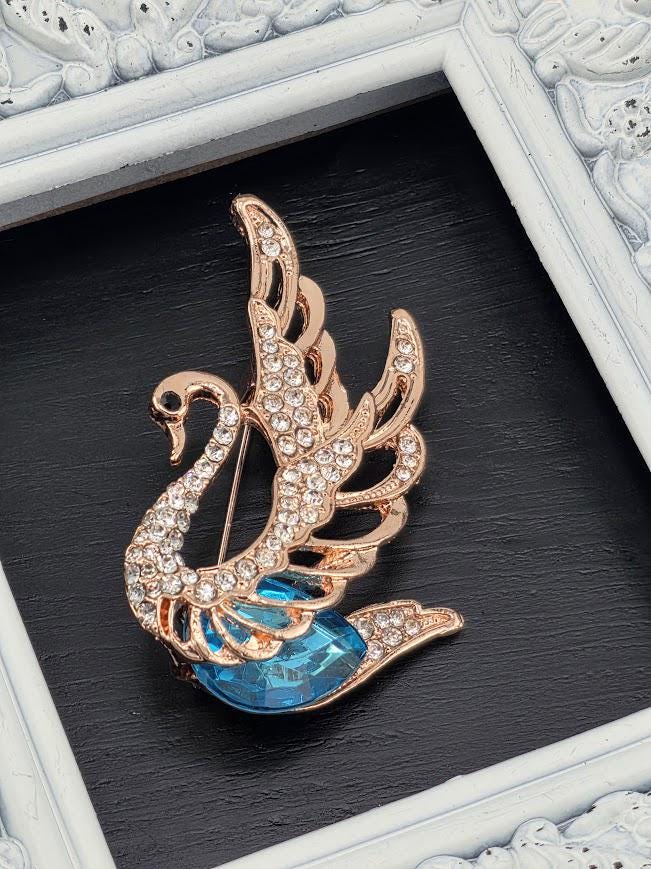 A decorative brooch in the shape of a swan, with a blue gemstone on its head, is displayed against a black background.