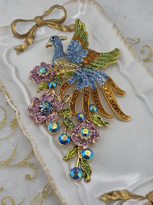 A decorative piece featuring a peacock with a blue body, green tail, and gold and blue accents, surrounded by colorful flowers and gemstones.