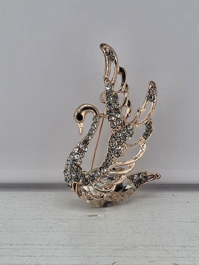 A decorative gold-colored brooch in the shape of a swan, adorned with sparkling rhinestones, is displayed within a white ornate frame against a dark background.