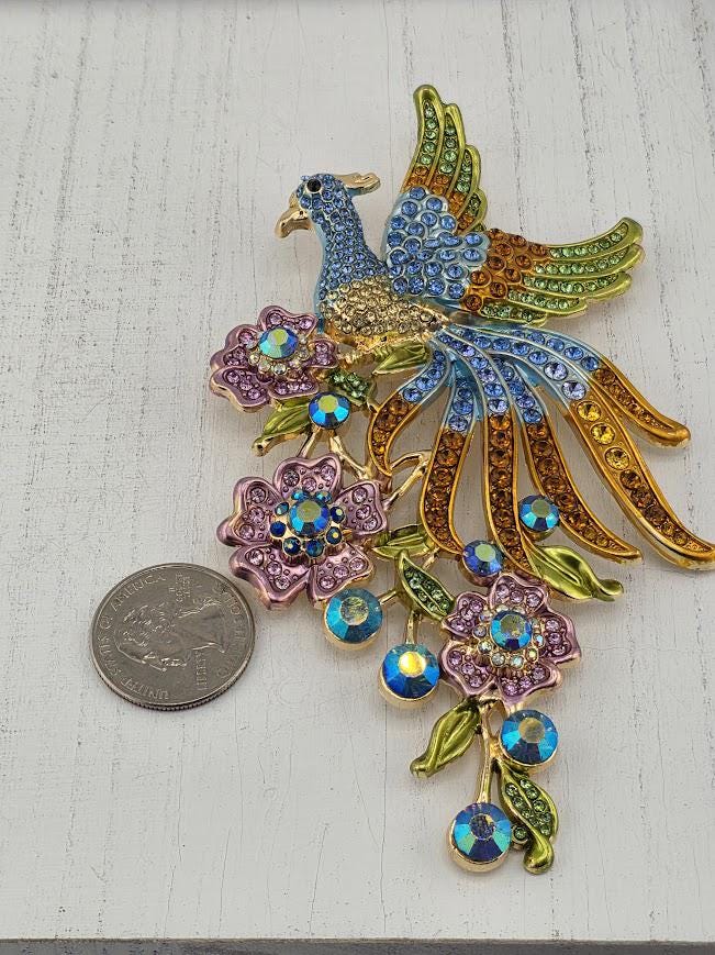 A decorative piece featuring a peacock with a blue body, green tail, and gold and blue accents, surrounded by colorful flowers and gemstones.