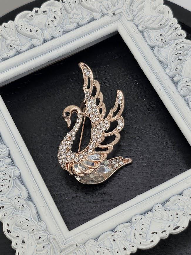 A decorative gold-colored brooch in the shape of a swan, adorned with sparkling rhinestones, is displayed within a white ornate frame against a dark background.