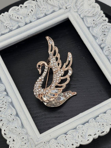 A decorative gold-colored brooch in the shape of a swan, adorned with sparkling rhinestones, is displayed within a white ornate frame against a dark background.