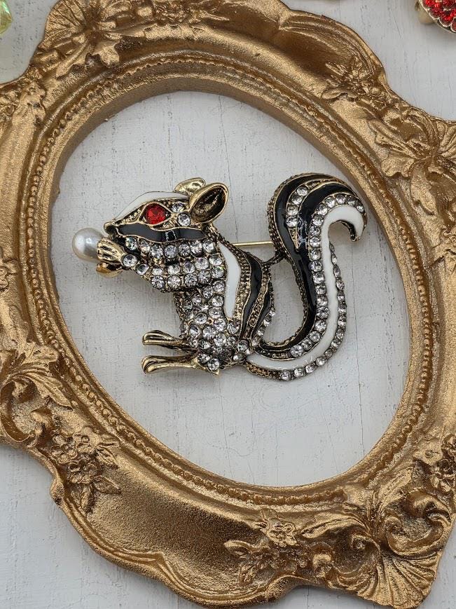 A decorative brooch in the shape of a skunk, adorned with rhinestones and pearls, is displayed within an ornate gold frame.