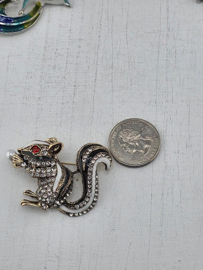 A decorative brooch in the shape of a skunk, adorned with rhinestones and a pearl, is placed next to a United States quarter on a white surface.