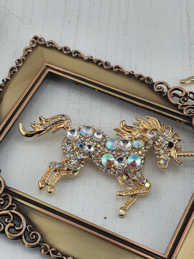 A decorative gold-colored brooch in the shape of a unicorn, adorned with numerous rhinestones and crystals, is displayed within an ornate gold frame.