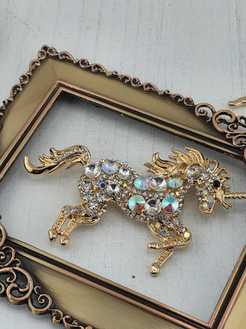 A decorative gold-colored brooch in the shape of a unicorn, adorned with numerous rhinestones and crystals, is displayed within an ornate gold frame.