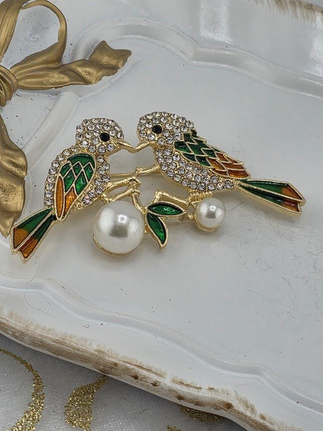 A decorative brooch featuring two colorful birds with gemstone accents and pearls.