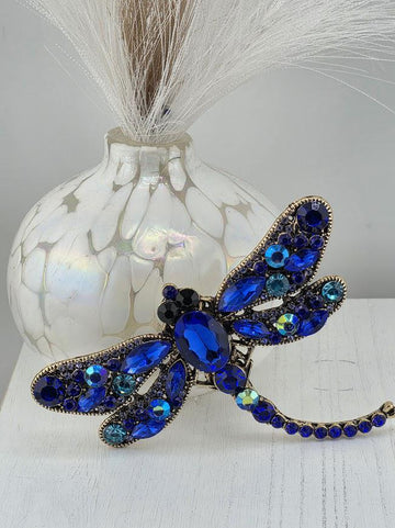 A decorative blue dragonfly brooch with blue gemstones and a white vase with white flowers in the background.