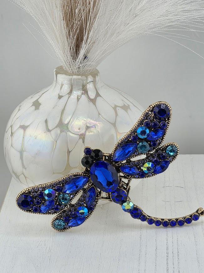 A decorative blue dragonfly brooch with blue gemstones and a white vase with white flowers in the background.