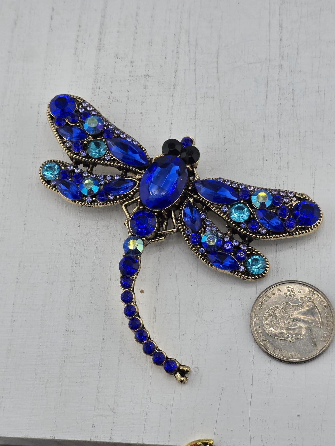 A decorative blue dragonfly brooch with blue gemstones and a white vase with white flowers in the background.