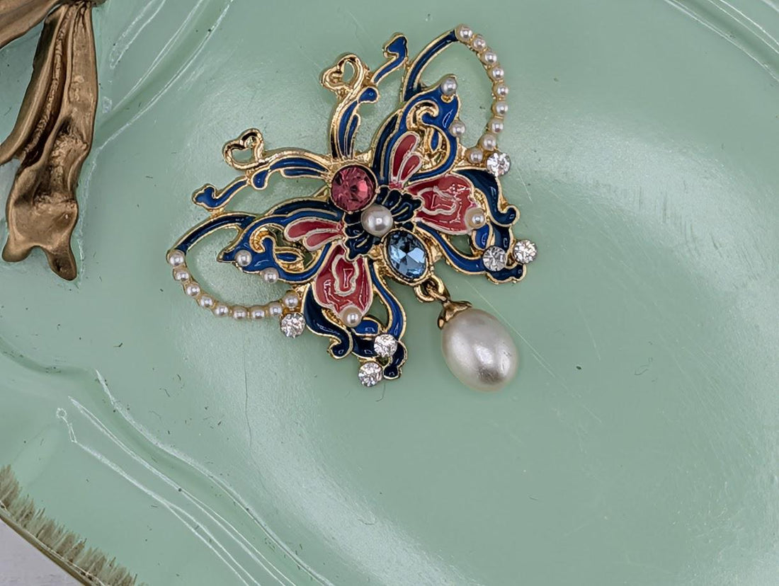 A decorative brooch with a pearl and gemstone accents, displayed in a white ornate frame.