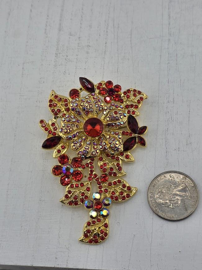 A decorative brooch with a large red gemstone surrounded by smaller red gemstones and leaves, placed on a white surface next to a coin.