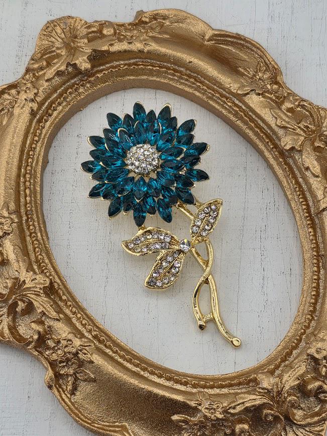 A decorative gold-framed brooch with a blue flower-shaped brooch and a leaf-shaped brooch is displayed on a white surface.
