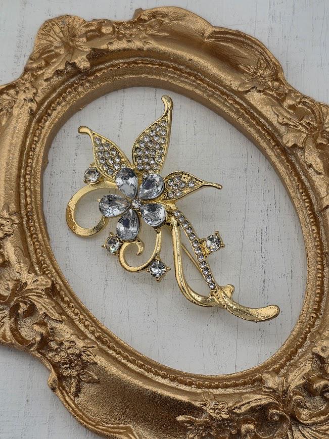 A decorative brooch with a floral design and sparkling gemstones is displayed in a white ornate frame.