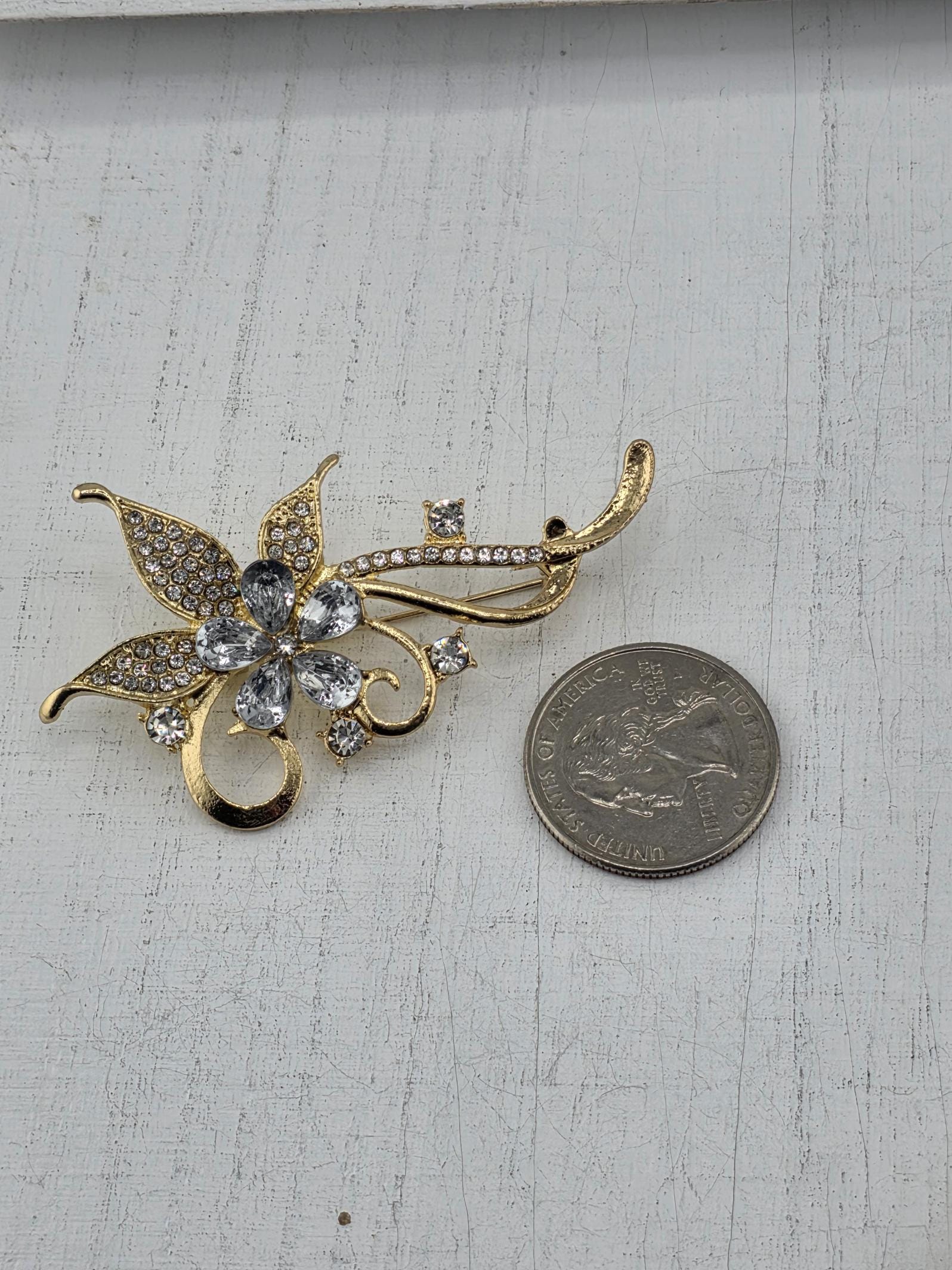 A decorative brooch with a flower-like design and sparkling gemstones is placed next to a United States quarter on a white wooden surface.