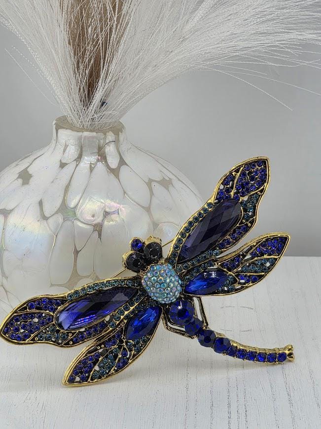 A decorative dragonfly brooch with blue and gold gemstones is displayed on a white surface, accompanied by a white vase with a feather-like decoration.