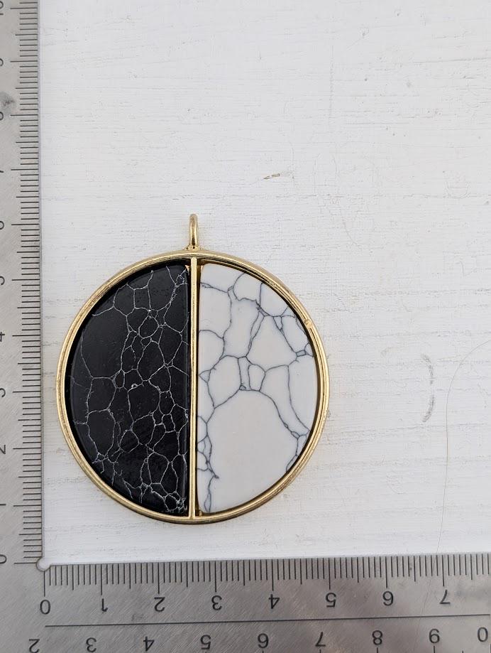 A gold-colored pendant with a black and white marble-like stone in the center, placed on a white surface next to a ruler for size reference.