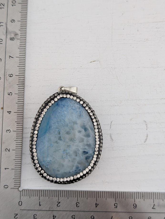 A blue geode pendant with a silver frame and rhinestones, placed on a ruler for size comparison.