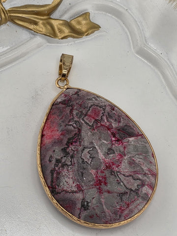 A large, oval-shaped pendant made of a dark-colored stone, possibly jade, with a gold-colored frame. The pendant is hanging from a gold-colored chain.