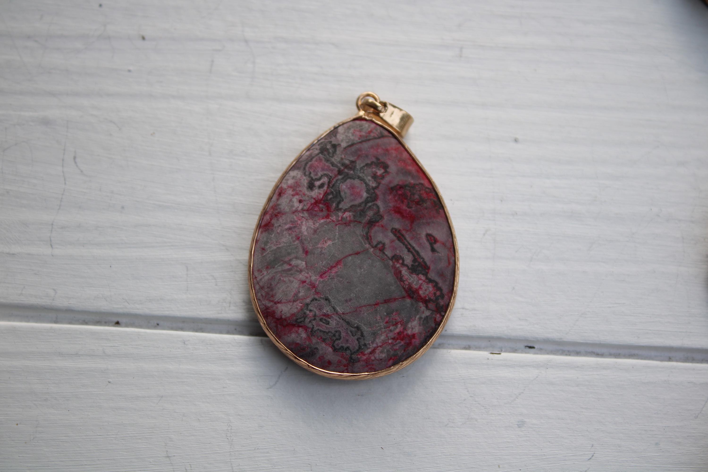 A pendant with a red and gray stone-like design, possibly a type of rock or gemstone, is displayed on a white wooden surface.