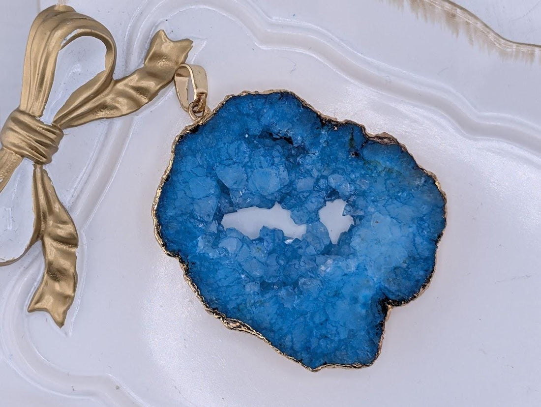 A close-up image of a blue geode pendant with a gold-colored metal frame, placed on a white surface.