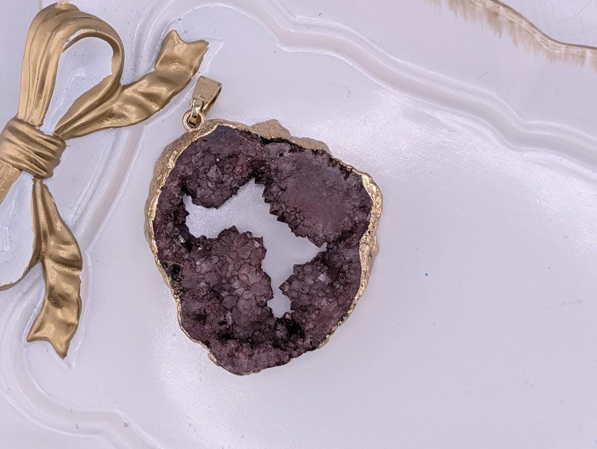 A large purple geode with a gold-colored pendant is displayed on a white surface, accompanied by a gold ribbon.