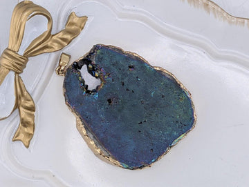 A large, blue-green geode pendant with a gold-colored border, placed on a white surface next to a gold ribbon.
