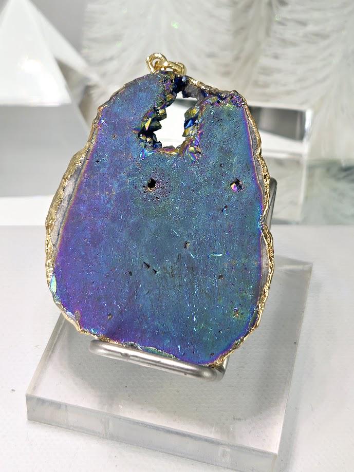 A large, blue-green geode pendant with a gold-colored border, placed on a white surface next to a gold ribbon.
