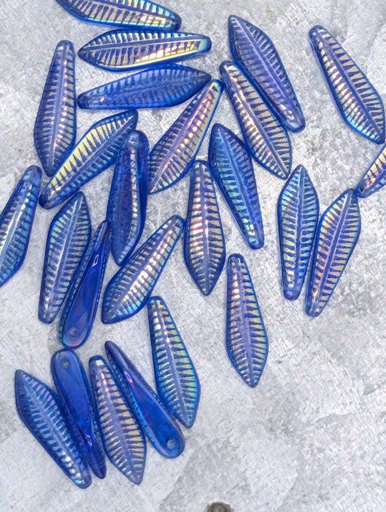 a collection of blue glass beads with a repeating pattern of leaf-like designs, scattered on a gray surface.