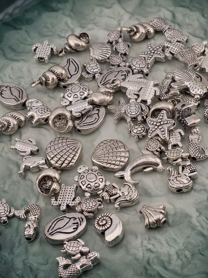 a collection of various silver-colored metal charms, including sea creatures such as fish, shells, and sea stars, as well as other metallic objects like coins and keys, scattered across a surface.