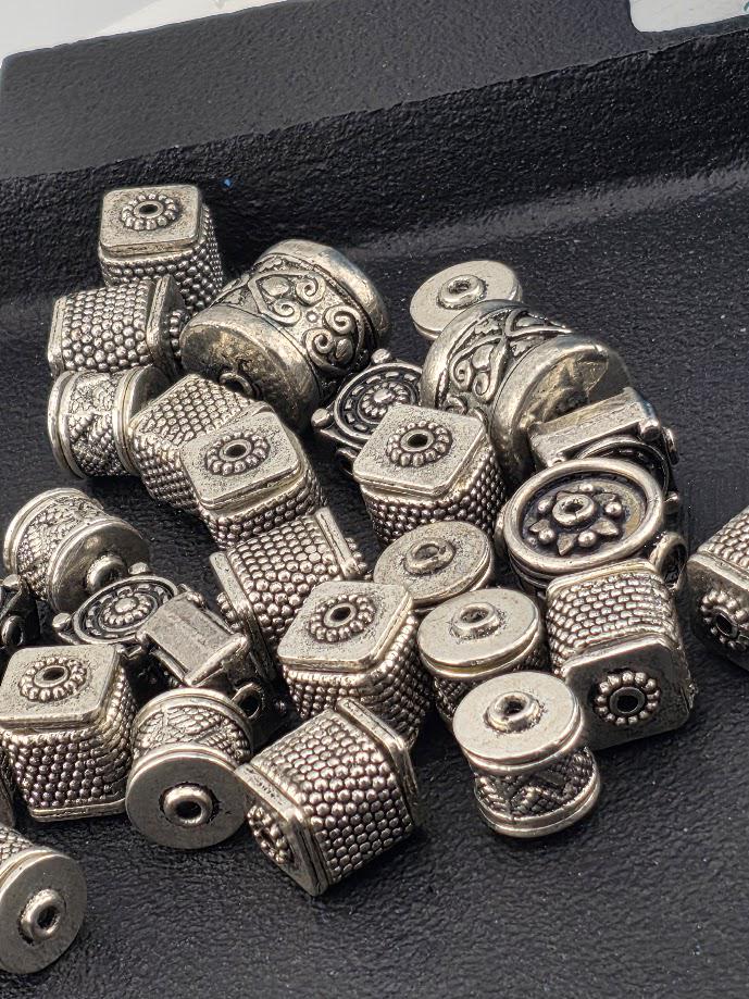 a collection of various metal beads and charms, including square and round shapes, scattered on a black surface.